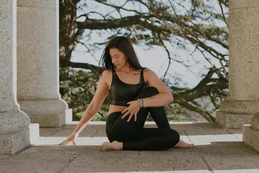 Woman practicing yoga outdoors near a stone structure, mid-twist pose.